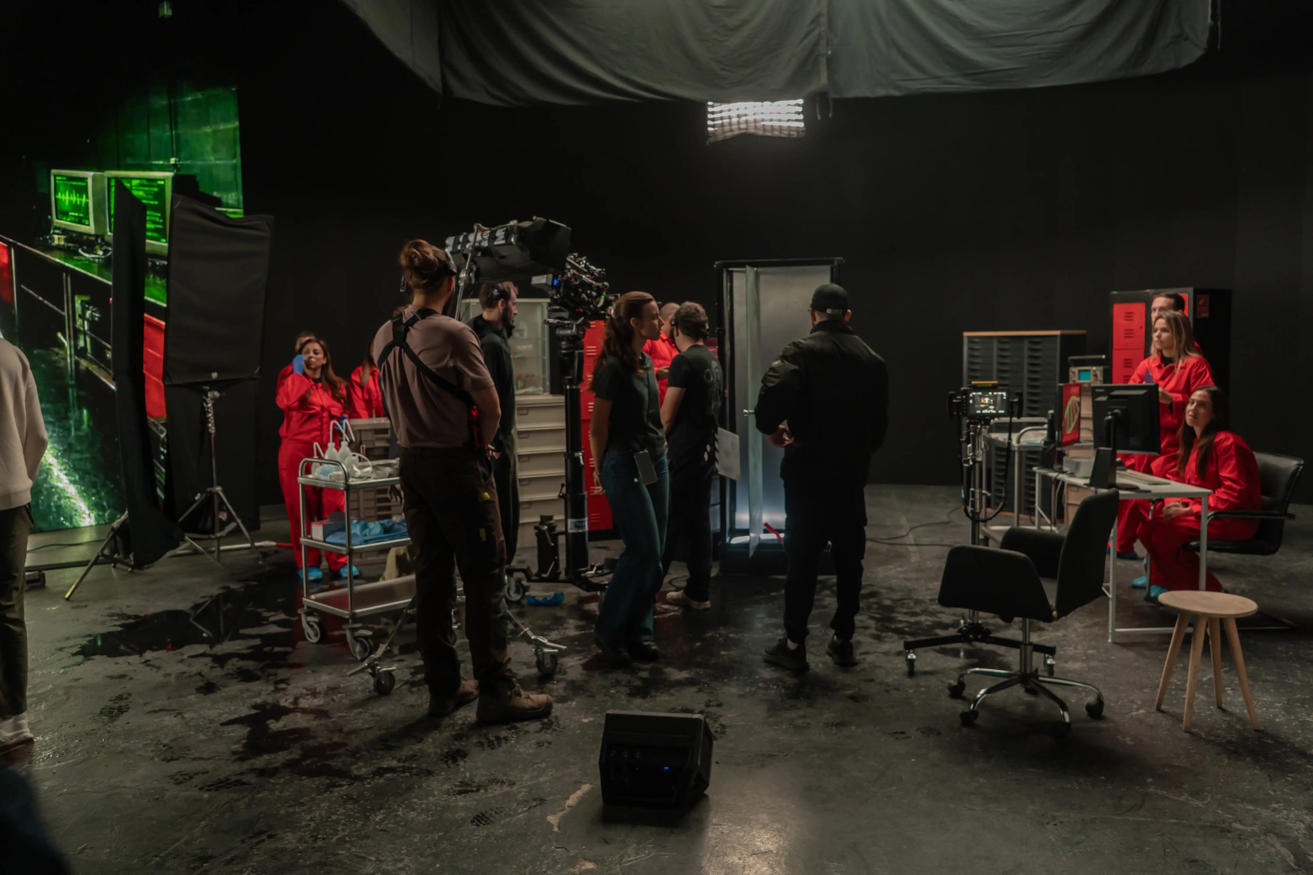 Équipe de tournage et figurants en combinaison rouge autour du caisson, devant le mur LED qui affiche le laboratoire généré par IA